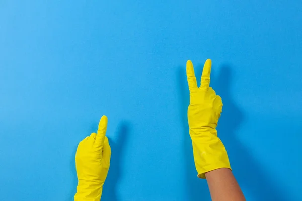 Mãos de mulher com luvas de borracha amarela fazendo um gesto que significa vitória e aponta para cima com o dedo, fundo azul — Fotografia de Stock