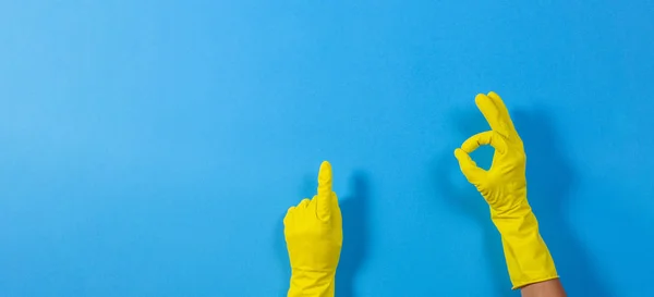 Mãos de mulher com luvas de borracha amarela fazendo um gesto que significa ok e aponta para cima com o dedo, fundo azul — Fotografia de Stock