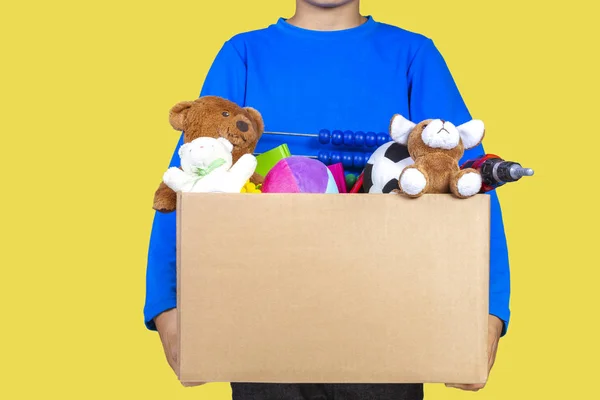 Concetto di donazione. Ragazzo in possesso di donare scatola con vestiti, libri, materiale scolastico e giocattoli, sfondo di colore giallo — Foto Stock