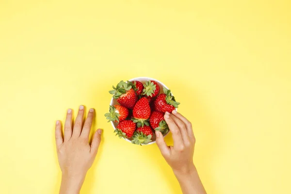 Kid hand taking strawberry from the bowl on yellow background