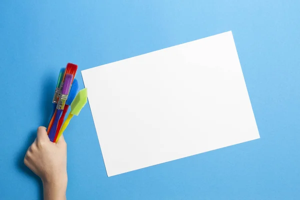 Mão de criança com pincéis coloridos e folha de papel branco em branco no fundo azul Fotografia De Stock