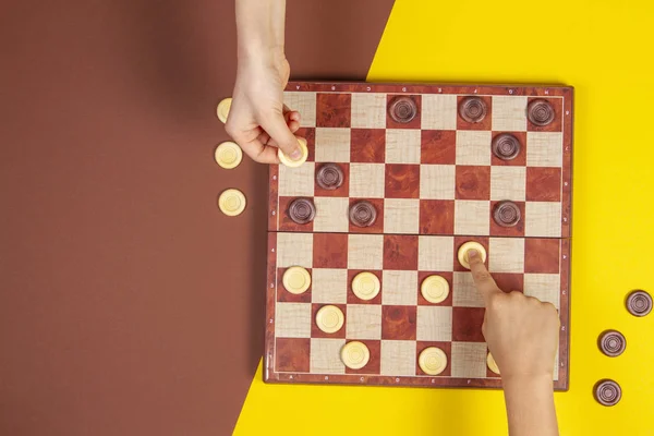 Child hands playing checkers on checker board game over yellow and blue background, top view — Stock Photo, Image