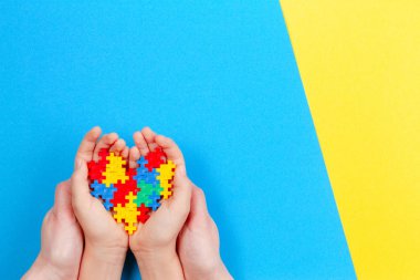 Adult and kid hands holding colorful heart on yellow and blue background. World autism awareness day concept clipart