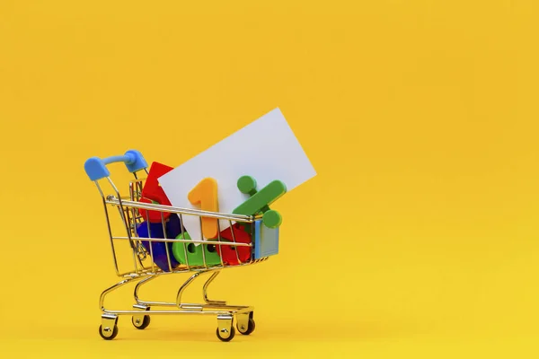 Carrinho de compras ou carrinho de supermercado cheio de números coloridos e cartão branco no fundo amarelo — Fotografia de Stock