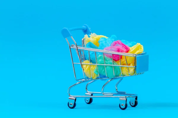 Mini carrinho de compras cheio de coloridos sacos de plástico de uso único no fundo azul — Fotografia de Stock