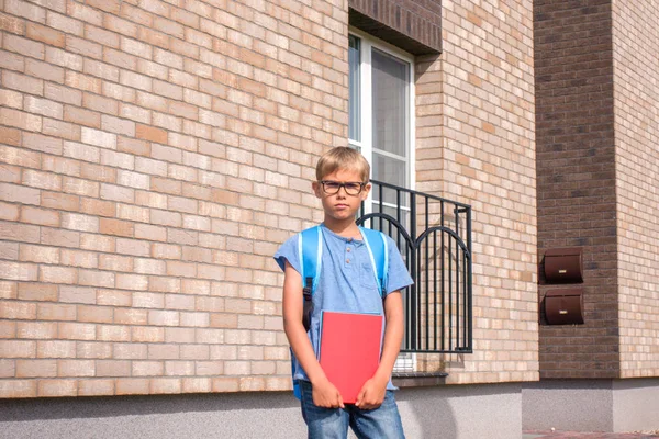 Chłopiec z czerwonym notebookiem i plecakiem stojący w pobliżu mieszkania mieszkalnego. Dziecko gotowe do szkoły — Zdjęcie stockowe