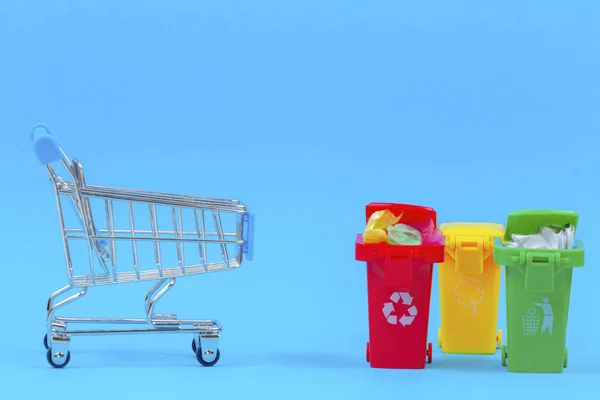 Mini carrinho de compras perto de recipientes de lixeiras no fundo azul — Fotografia de Stock