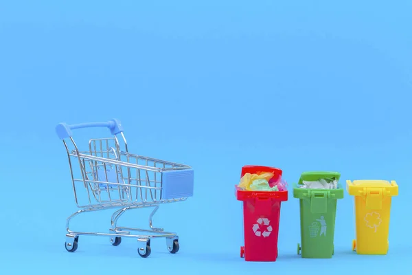 Troli belanja mini di dekat tempat sampah daur ulang berisi wadah dengan latar belakang biru — Stok Foto