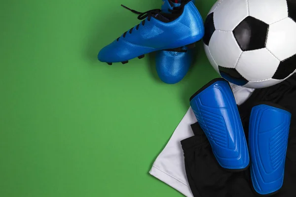 Soccer ball, blue boots, cleats, white t-shirt and black shorts on green background. Flat lay, top view