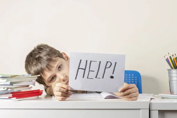 Triste garçon frustré fatigué assis à la table avec de nombreux livres et tenant du papier avec mot Aide. Difficultés d'apprentissage, concept éducatif . — Photo