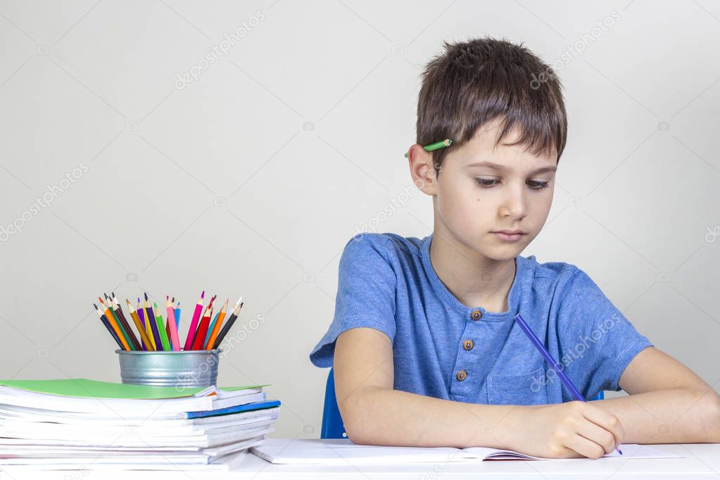 Kid doing homework at the table. Focused boy with pencil behind his ear writing with pencil