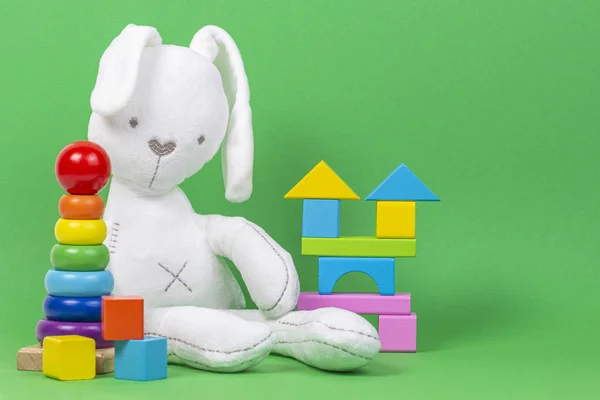 White plush toy rabbit, baby stacking rings pyramid and colorful wooden blocks on light green background