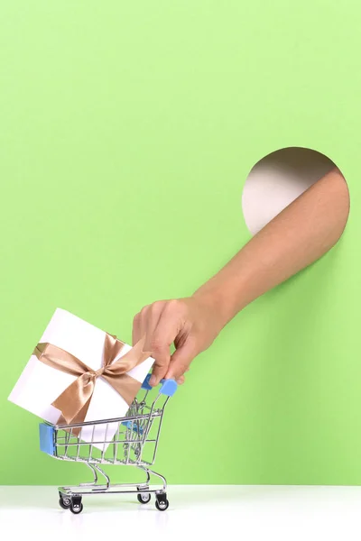 밝은 녹색 배경에 구멍을 통해 선물 선물 상자와 미니 쇼핑 트롤리를 만지는 손 — 스톡 사진