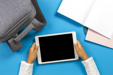 Açık mavi arka plan üzerinde tablet bilgisayar, sırt çantası ve renkli kağıt dizüstü bilgisayar ile çocuk eller için üst görünüm