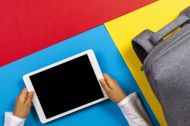 Renkli arka planda tablet bilgisayar ve gri sırt çantası ile çocuk ellerine üst görünüm