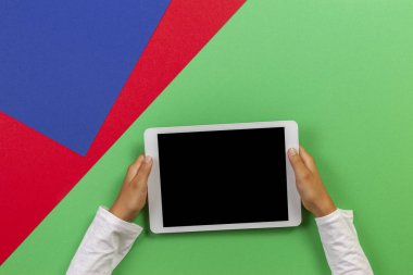 Açık yeşil, mavi ve kırmızı arka plan üzerinde beyaz tablet bilgisayar tutan çocuk elleri