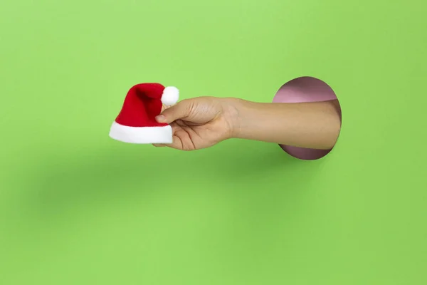 Mão através de buraco em luz verde fundo segurando pequeno vermelho Natal Papai Noel chapéu — Fotografia de Stock