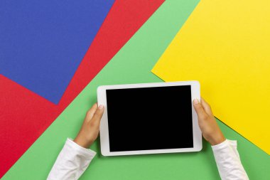 Açık yeşil, mavi, sarı ve kırmızı arka planda beyaz tablet bilgisayarı tutan çocuk elleri
