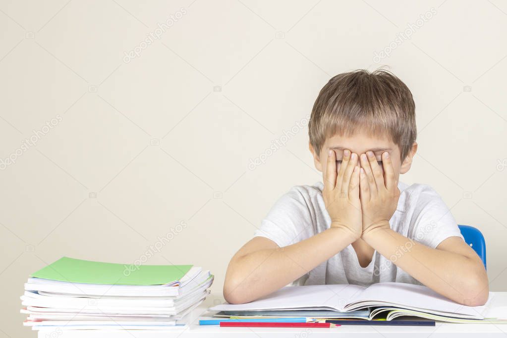 Sad tired upset schoolboy covering face with his hands sitting near table with pile of school books and notebooks