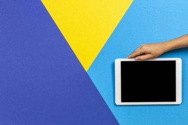 Açık mavi, sarı ve mavi arka planda beyaz tablet bilgisayar tutan çocuk el