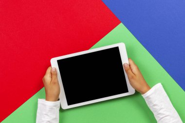 Açık yeşil, mavi ve kırmızı arka plan üzerinde beyaz tablet bilgisayar tutan çocuk elleri
