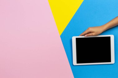 Açık mavi, sarı ve pastel pembe arka plan üzerinde beyaz tablet bilgisayar tutan çocuk el