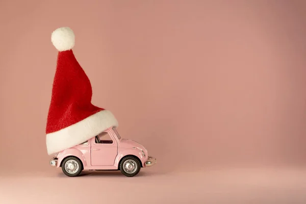 Christmas background. Little retro toy model car with small red Christmas Santa Claus hat on pastel pink background