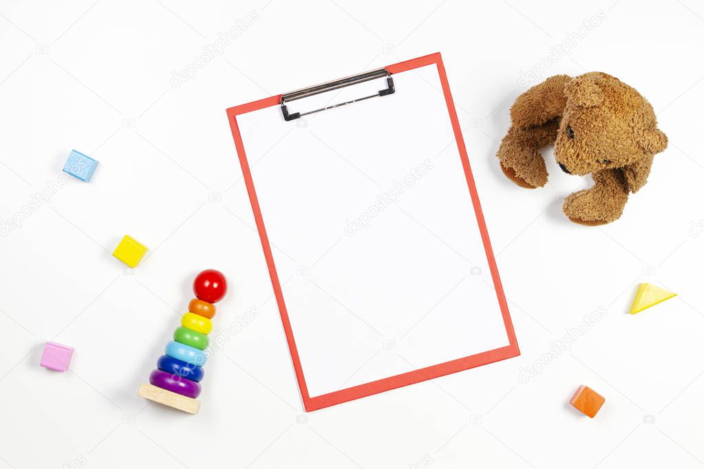 Colorful baby kid toys and red clipboard with blank sheet of paper on white background. Top view