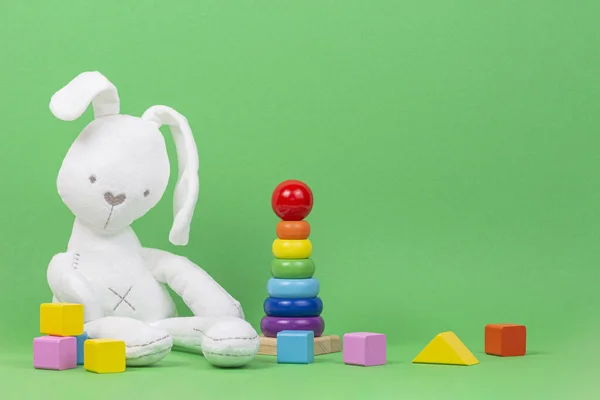 White plush toy rabbit, baby stacking rings pyramid and colorful wooden blocks on light green background