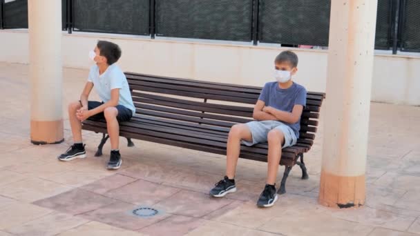 Діти з медичними захисними масками сидять на лавці на відкритому повітрі. Хлопці тримають соціальне дистанціювання, щоб запобігти поширенню вірусу — стокове відео