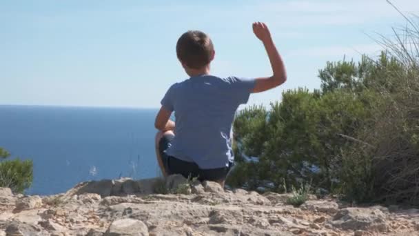 Один грустный мальчик сидит на земле и бросает камни в воду в солнечный день — стоковое видео