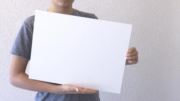 Мальчик держит в руках пустой белый макет плаката. Серая стена на заднем плане. Закрыть — стоковое видео
