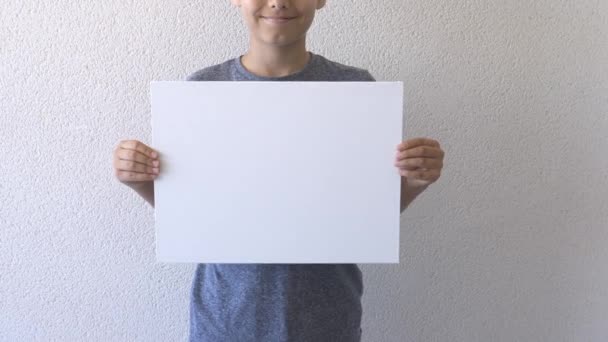 Хлопчик тримає в руках і показує порожній білий наклеп на дошці. Сіра стіна на фоні. крупним планом — стокове відео