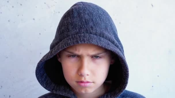 Ongelukkige verdrietige boos tiener jongen in een kap staan in de buurt van grijze muur en kijken naar camera — Stockvideo