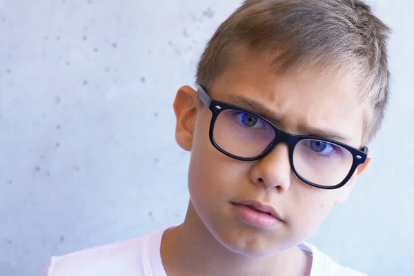 Niño preocupado inteligente con ojos azules gafas de bloqueo de luz mirando a la cámara. Grunge fondo de pared gris —  Fotos de Stock