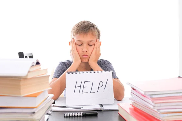 Fáradt, frusztrált fiú ül az asztalnál, a házi feladatát egy halom könyv között csinálja. A Word Help nyílt füzetre van írva. — Stock Fotó