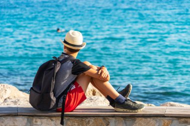 Güneş fötr şapkalı ve koruyucu maskeli çocuk deniz kenarındaki bankta oturuyor. Seyahat, tatil, tatil konsepti