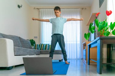 Dizüstü bilgisayarı olan çocuk spor egzersizleri yapıyor, evde yoga yapıyor. Spor, sağlıklı yaşam tarzı, çocuklar için aktif eğlence.