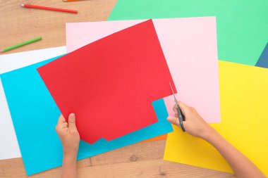 Çocuk elleri ahşap masada renkli kağıtları makasla kesiyor. Üst görünüm