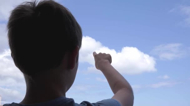 Chlapec ukazuje rukou na modrou oblohu a prstem něco kreslí ve vzduchu — Stock video