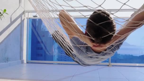 Child laying and relaxing in hammock hanging on balcony of apartment. Back view. Kid swinging and dreaming, thinking — Stock Video