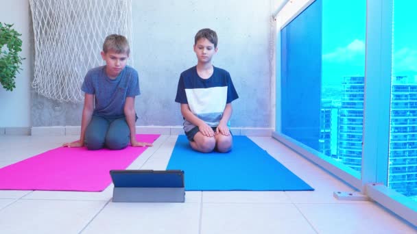 Barn som motionerar, stretchar på yogamattan medan de tittar på videolektioner online på surfplatta på balkong. Sport, hälsosam livsstil, aktiv fritid i hemmet — Stockvideo