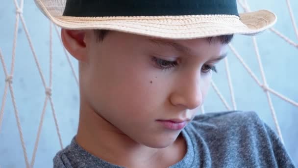 サンフェドーラの帽子をかぶった悲しい子供はハンモックに座って何かを考えています。閉めろ。4kビデオ — ストック動画