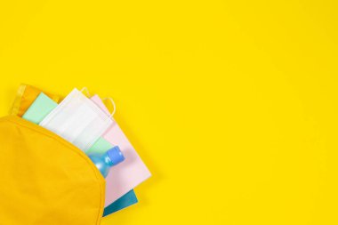 Eğitim, öğrenme, salgın sırasında okula dönme. Sarı arka planda not defterleri, okul gereçleri, el dezenfektanı ve koruyucu sağlık maskesi olan sırt çantalarını açmak için en iyi manzara