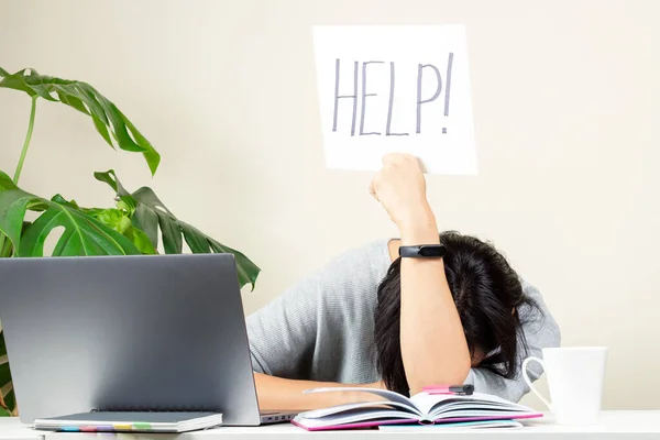 Стомлена жінка сидить за столом і тримає карту словом "Допоможіть". Працює в Інтернеті, дистанційне навчання з дому. — стокове фото