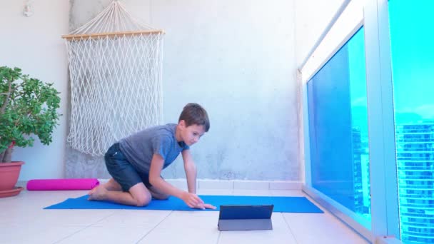 Dzieciak robi ćwiczenia sportowe, robi ćwiczenia deski na macie do jogi podczas oglądania lekcji wideo online na tablecie na balkonie. Sport, zdrowy tryb życia, aktywny wypoczynek w domu — Wideo stockowe
