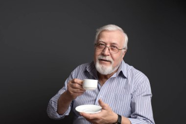 İyi huylu, güler yüzlü, gri arka planda beyaz bir bardak tutan ve çerçeveye bakan bir adam.