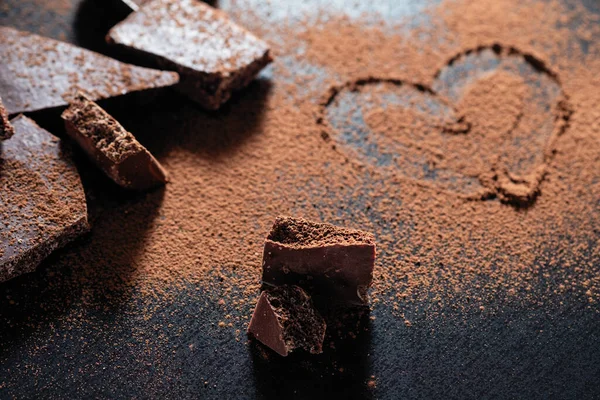 broken chocolate bar on a black background, cocoa powder painted heart