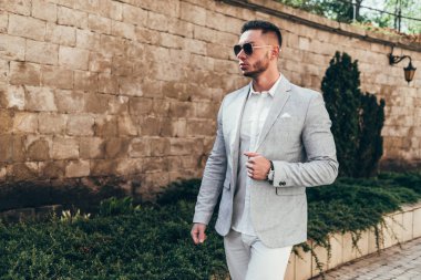 Sakallı ve güneş ışığı altında sokakta şık takım elbise poz içinde gözlüklü genç adam