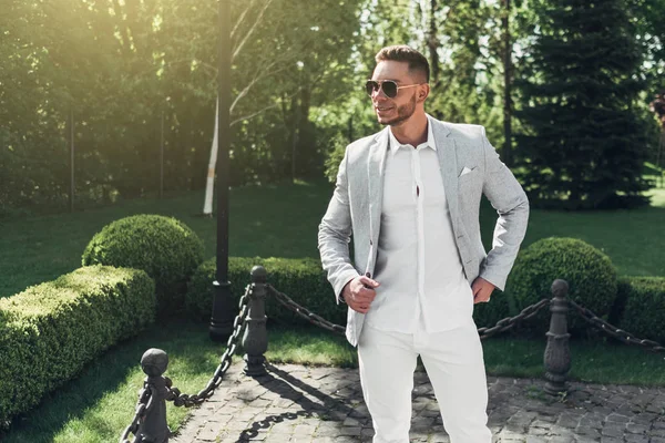 Sakallı ve güneş ışığı altında sokakta şık takım elbise poz içinde gözlüklü genç adam